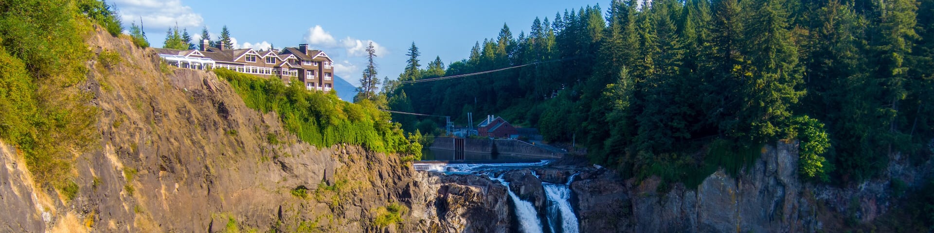 A waterfall cascading down a rocky cliff into a river, surrounded by dense forest and a building on a hill.