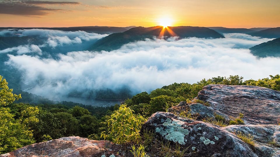 A sunrise over a mountainous landscape with clouds below and rocky outcrops in the foreground.