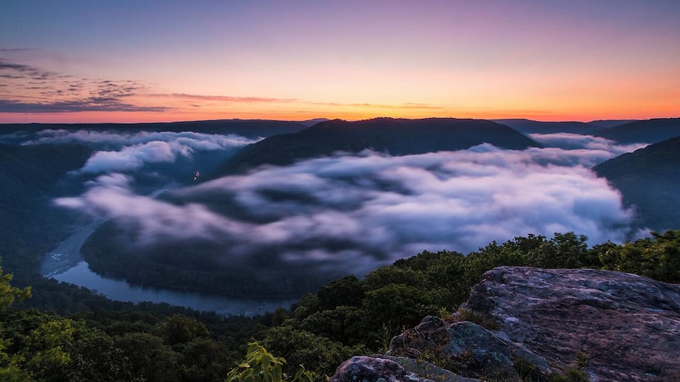 A mountainous landscape at sunset with a river winding through a valley, surrounded by clouds and dense forest.