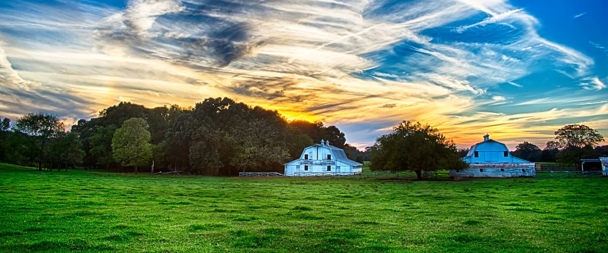 A rural landscape with a blue barn, a white church, and a field under a dramatic sky with clouds.