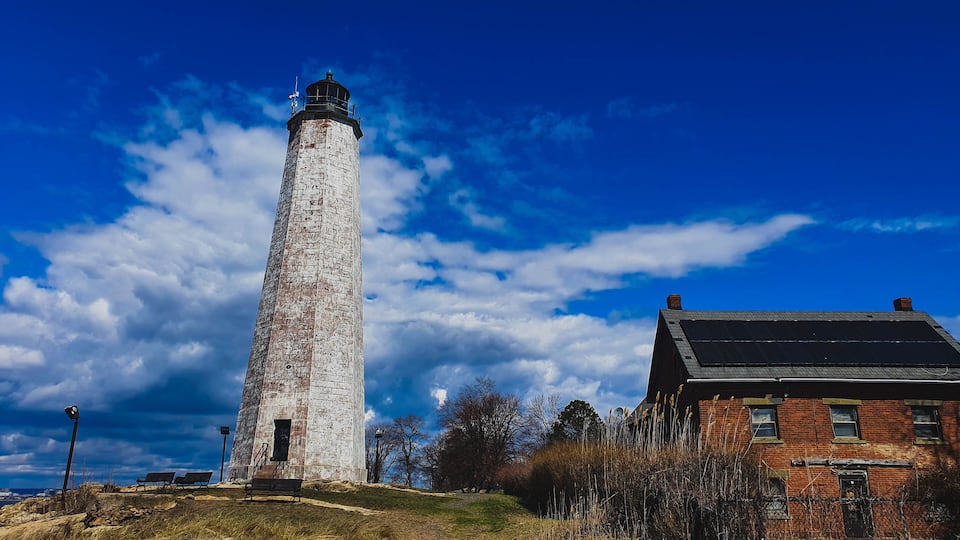 A tall white lighthouse with a black top, a brick building with a dark roof, and a clear blue sky with scattered clouds.