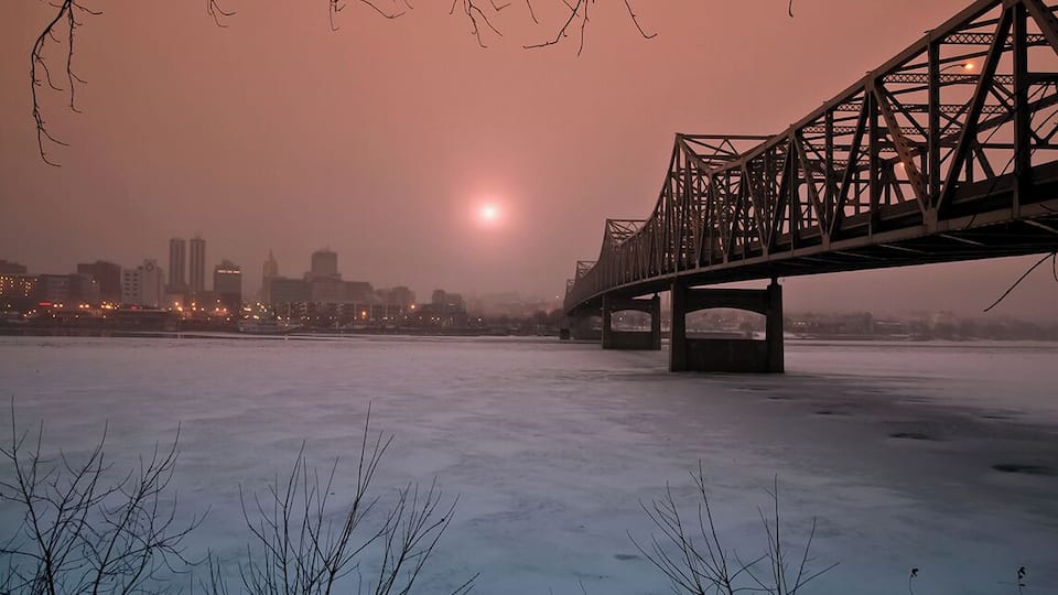 A bridge over a frozen body of water with a city skyline in the background.