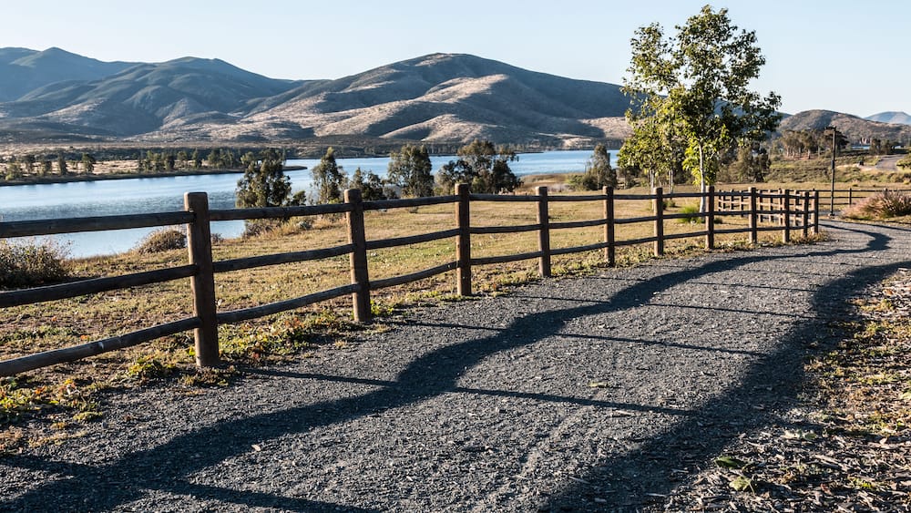 Un chemin de gravier bordé d’une clôture en bois, un lac et des montagnes en arrière-plan.