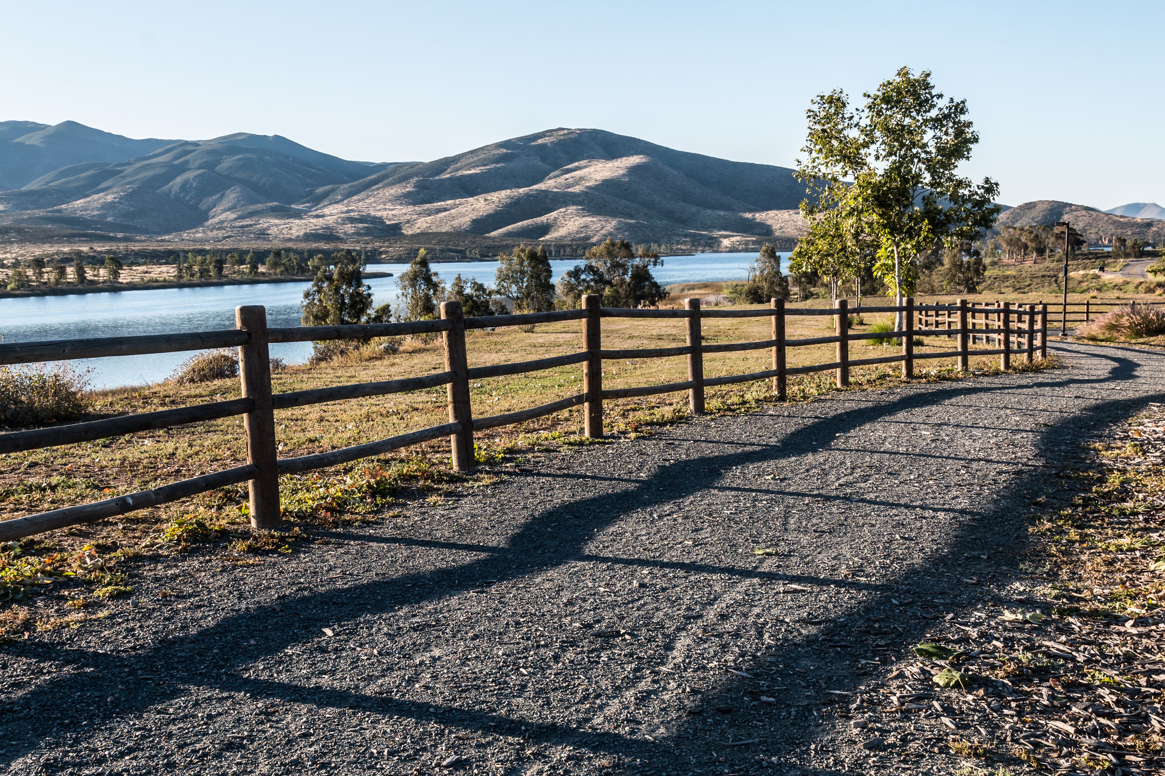 A gravel path with a wooden fence, a lake, and mountains in the background.