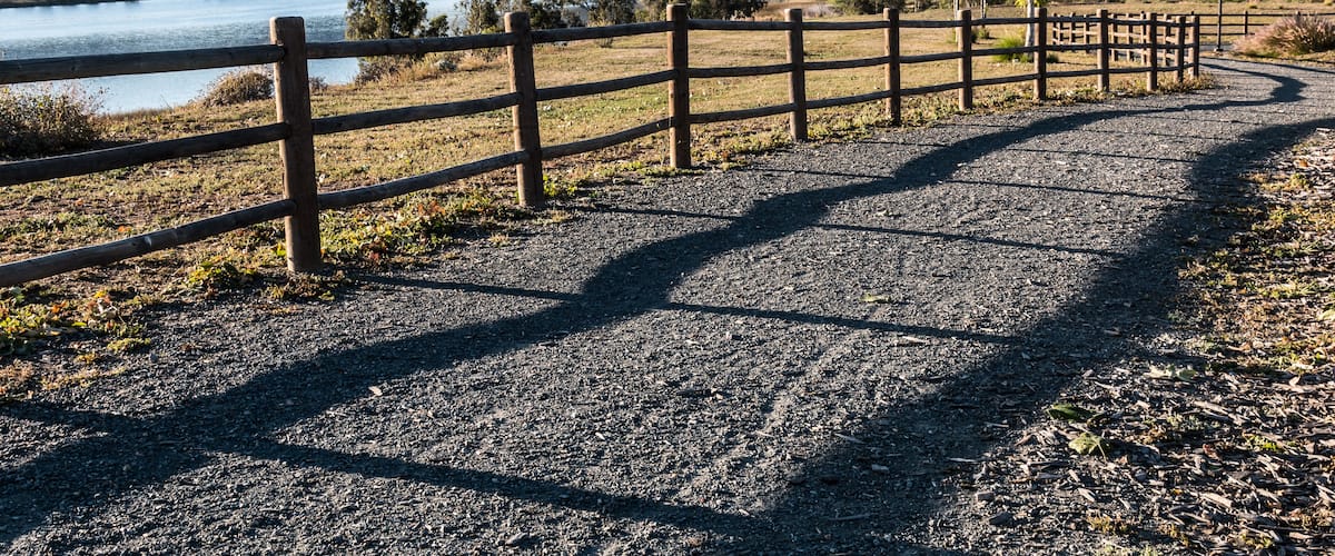 A gravel path with a wooden fence, a lake, and mountains in the background.