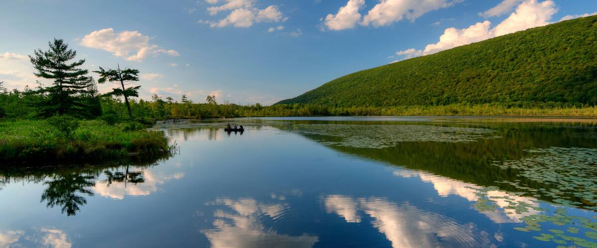 A calm lake with reflections of clouds and trees, surrounded by a lush green hillside.