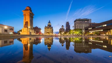 Paisagem urbana com um monumento imponente refletido em águas calmas, edifícios históricos e arquitetura moderna sob o céu crepuscular.