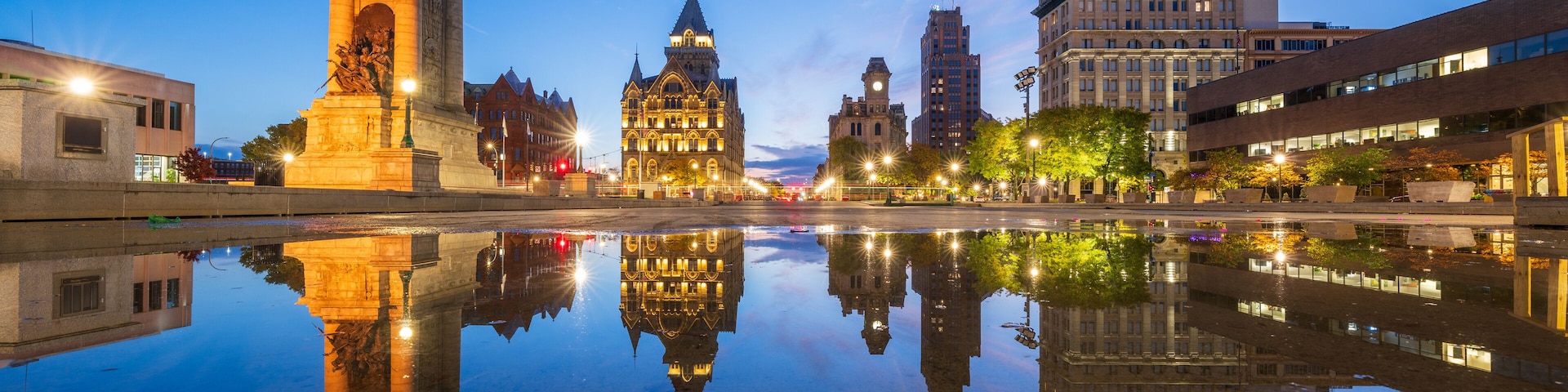 Un paisaje urbano con un monumento destacado reflejado en aguas tranquilas, edificios históricos y arquitectura moderna bajo un cielo crepuscular.