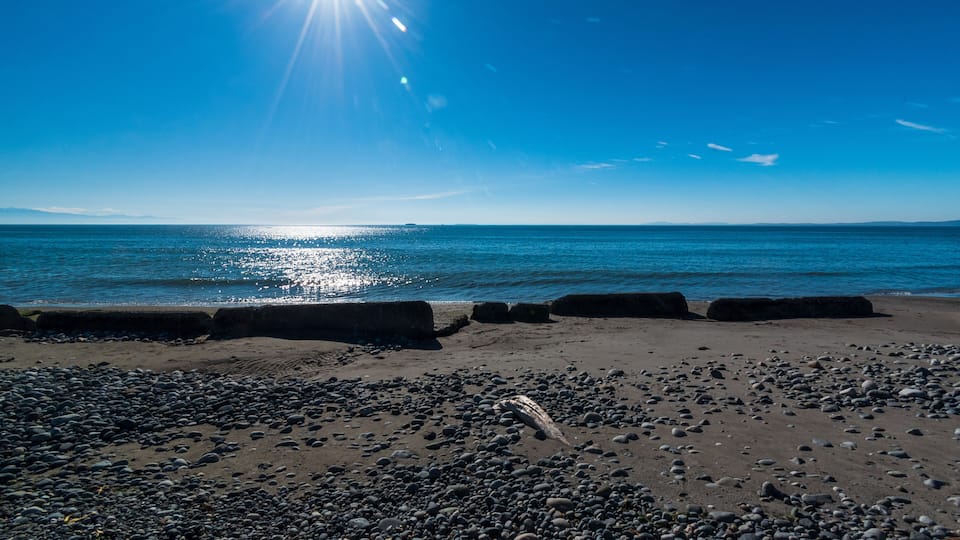 A beach with pebbles, calm water, and a clear sky.
