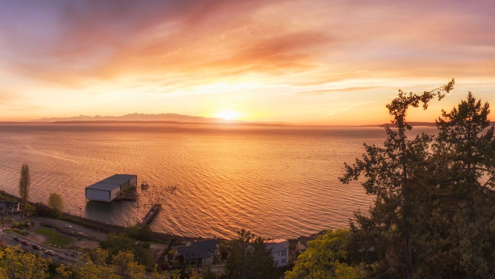 Paisagem costeira com o pôr do sol sobre a água, um cais e árvores ao redor.
