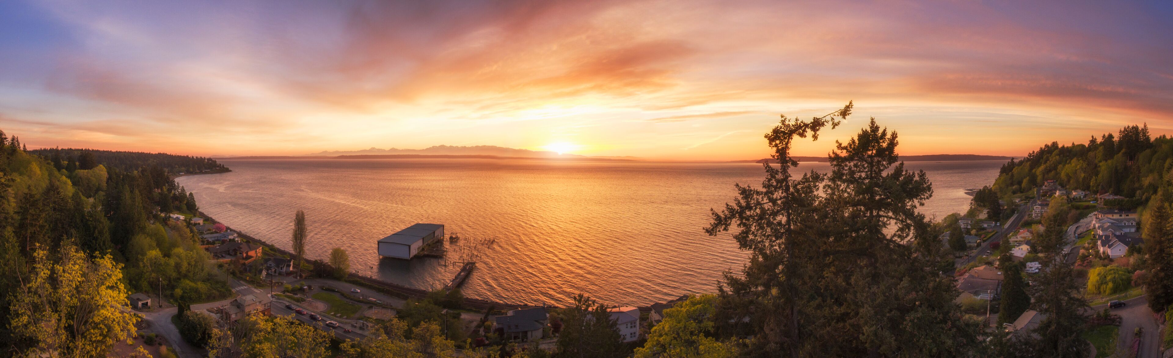 A coastal landscape with a sunset over the water, a dock, and surrounding trees.