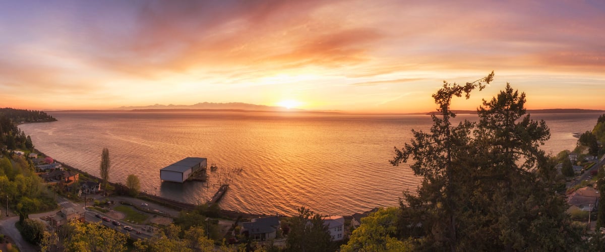A coastal landscape with a sunset over the water, a dock, and surrounding trees.