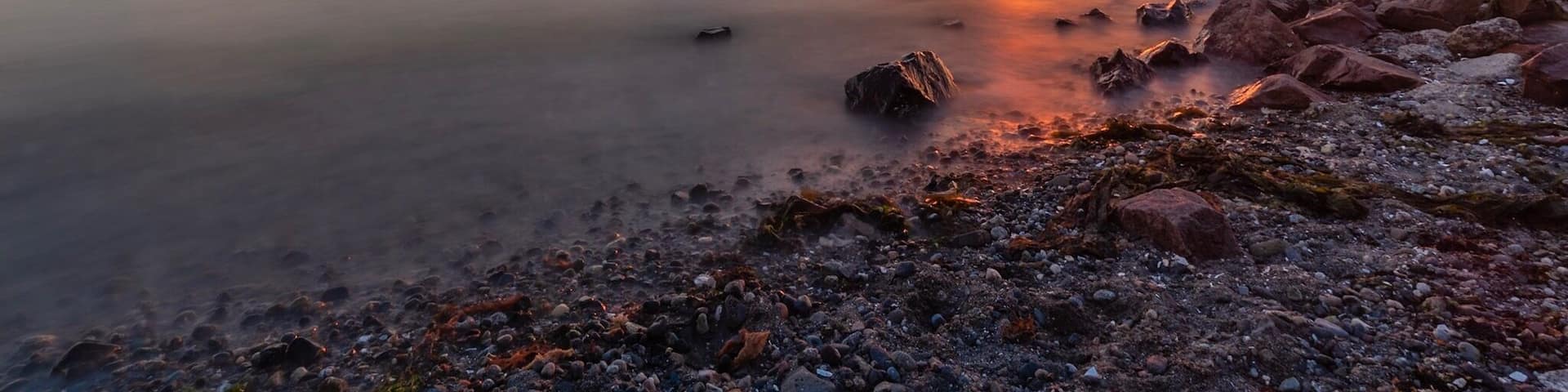 A sunset over a calm lake with rocky shores and distant mountains.