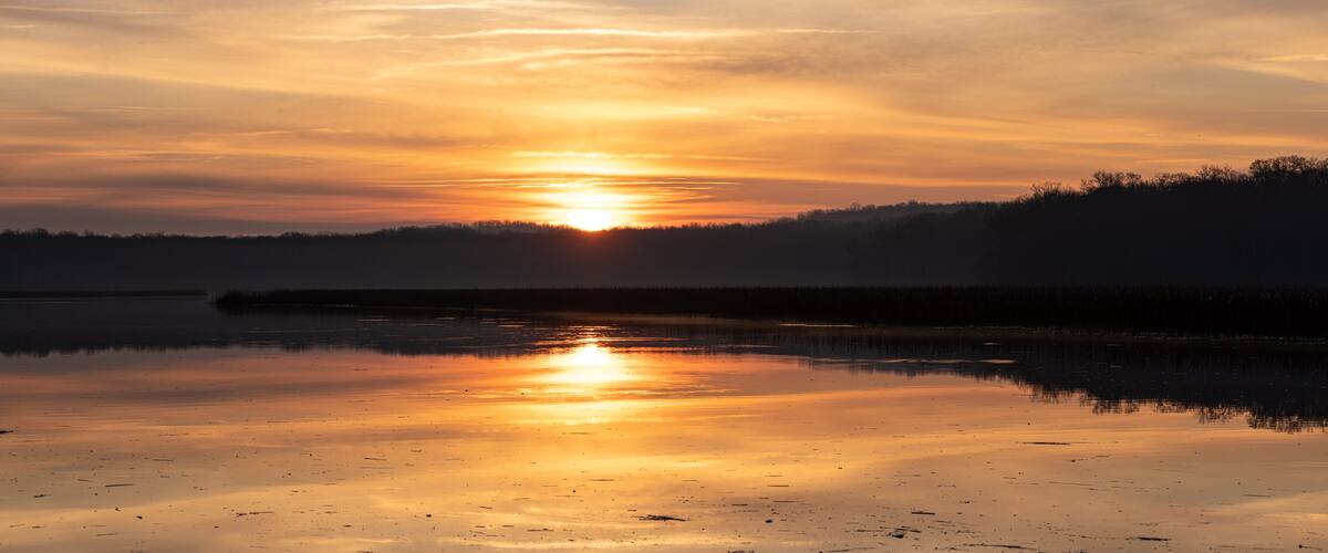 A sunset over a calm lake with reflections of the sun, surrounded by a forested area.