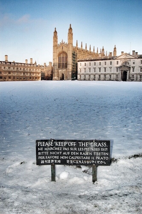 A snow-covered area with a sign in multiple languages asking to keep off the grass.