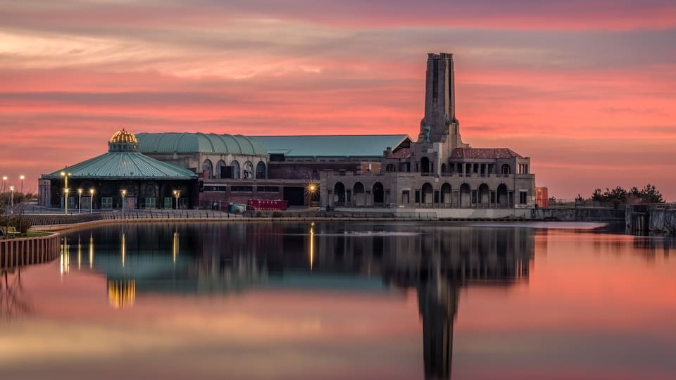 A historic building with a green dome and a tall tower, reflecting in a calm body of water during sunset.