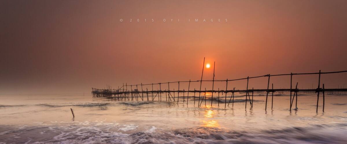 A beach at sunset with waves crashing onto the shore and a wooden pier extending into the water.