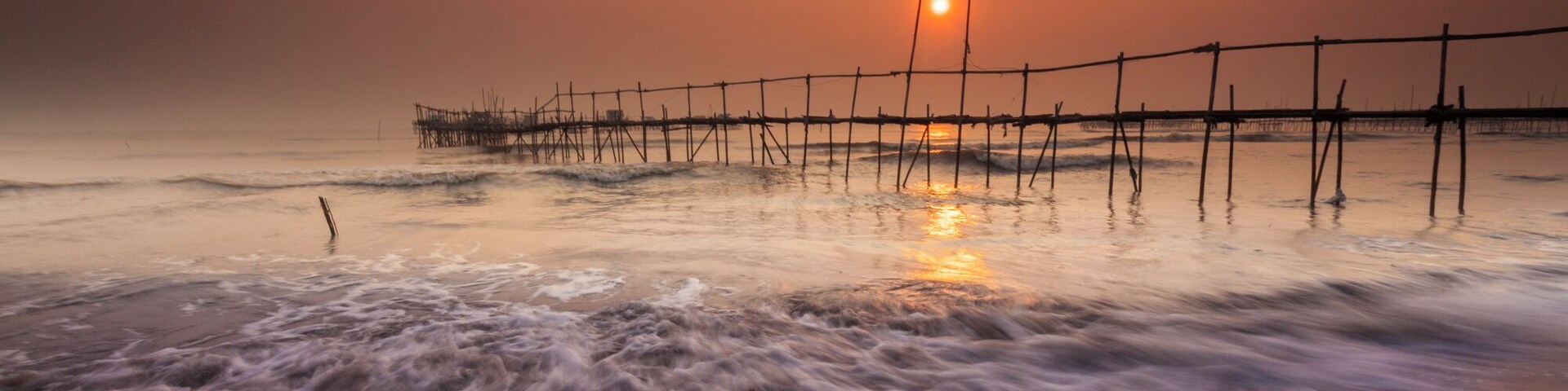 A beach at sunset with waves crashing onto the shore and a wooden pier extending into the water.