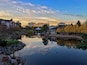 Escena serena junto al lago con follaje otoñal, un pabellón y edificios modernos al fondo.