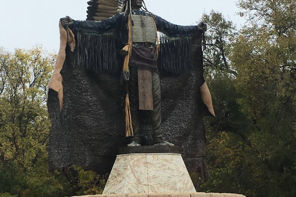 Una estatua de nativo americano con tocado emplumado, sobre un pedestal de piedra con una placa que dice 'Touch the Clouds'.