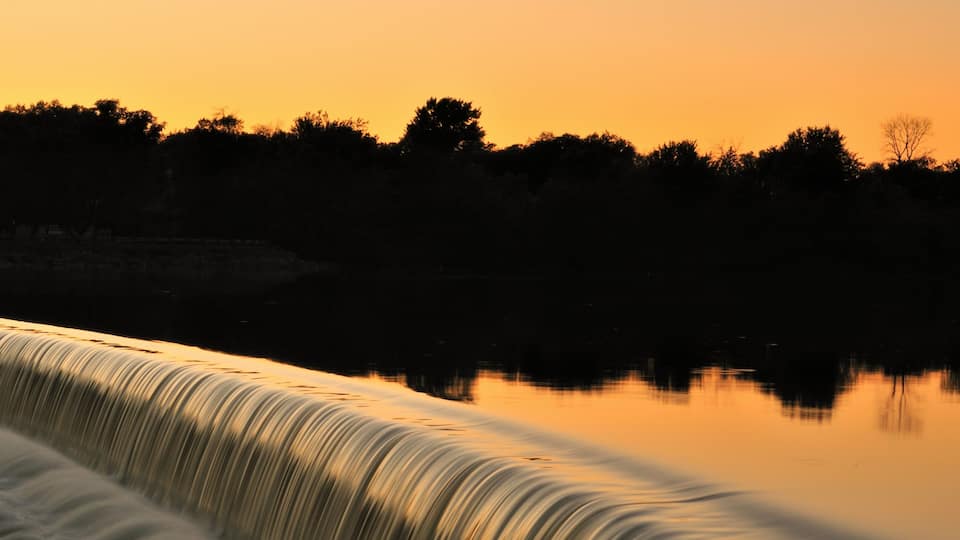Tranquil water turns to white water as it flows over a dam in the Fox River in South Elgin, Illinois while reflecting the the glow from the setting sun beyond the trees on the far bank.