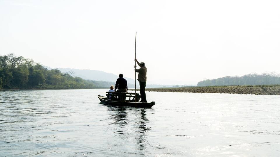 A calm river with a boat carrying two people, one standing and one seated, and a person fishing on the shore.