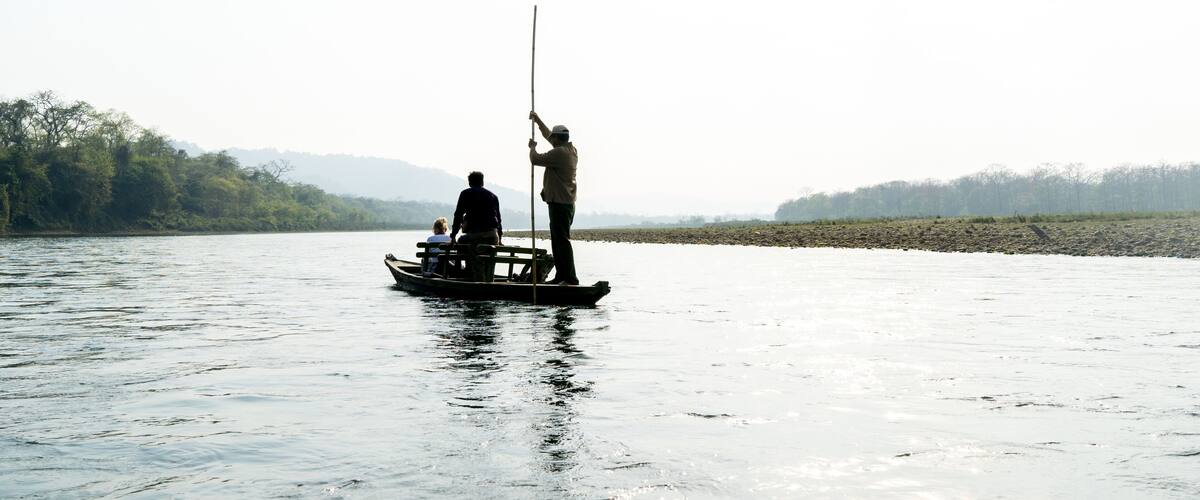 A calm river with a boat carrying two people, one standing and one seated, and a person fishing on the shore.