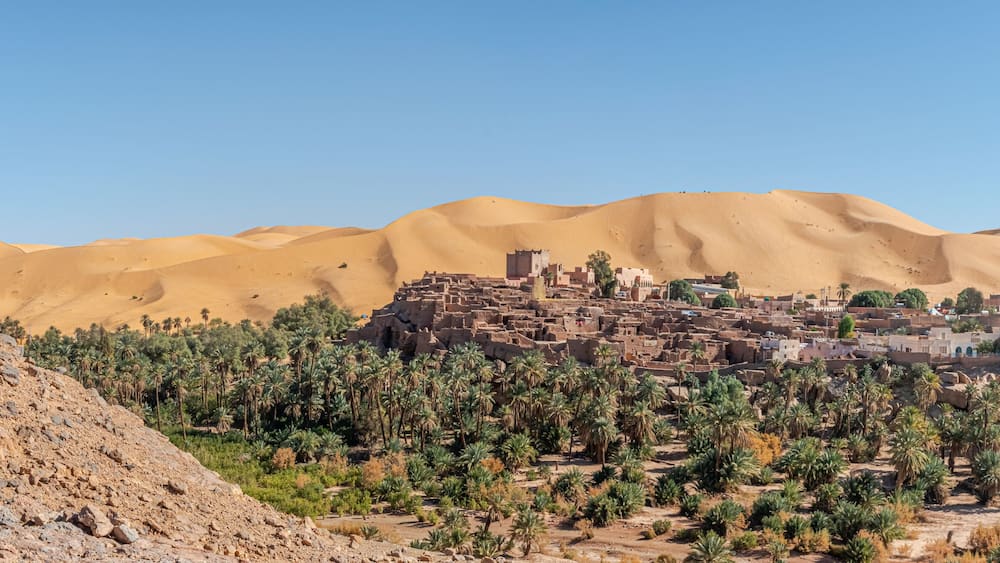 Panoramic view of Taghit in Bechar. Algerian Sahara desert oasis palm trees, trees, sand dunes and ksar old dry stone buildings. Aerial view from rocky mountain Djebel Baroun in foreground.