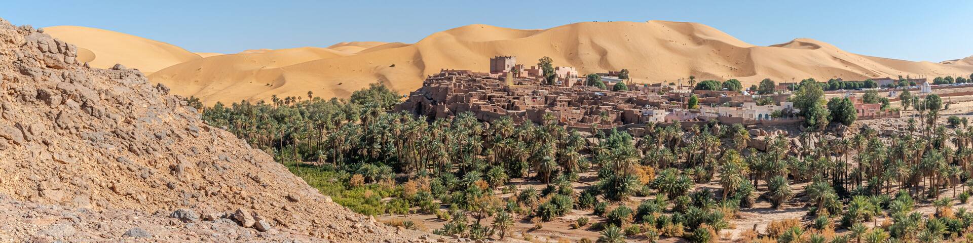 Panoramic view of Taghit in Bechar. Algerian Sahara desert oasis palm trees, trees, sand dunes and ksar old dry stone buildings. Aerial view from rocky mountain Djebel Baroun in foreground.
