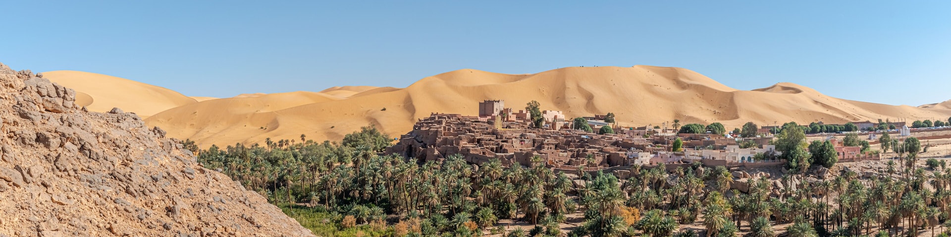 Panoramic view of Taghit in Bechar. Algerian Sahara desert oasis palm trees, trees, sand dunes and ksar old dry stone buildings. Aerial view from rocky mountain Djebel Baroun in foreground.