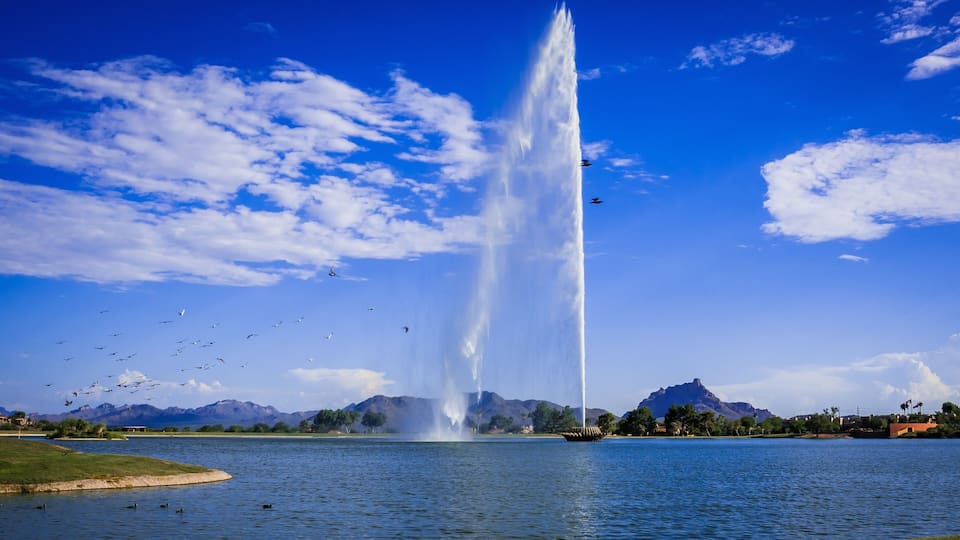 A fountain jetting water into the sky, surrounded by a lake, mountains, and a clear blue sky.