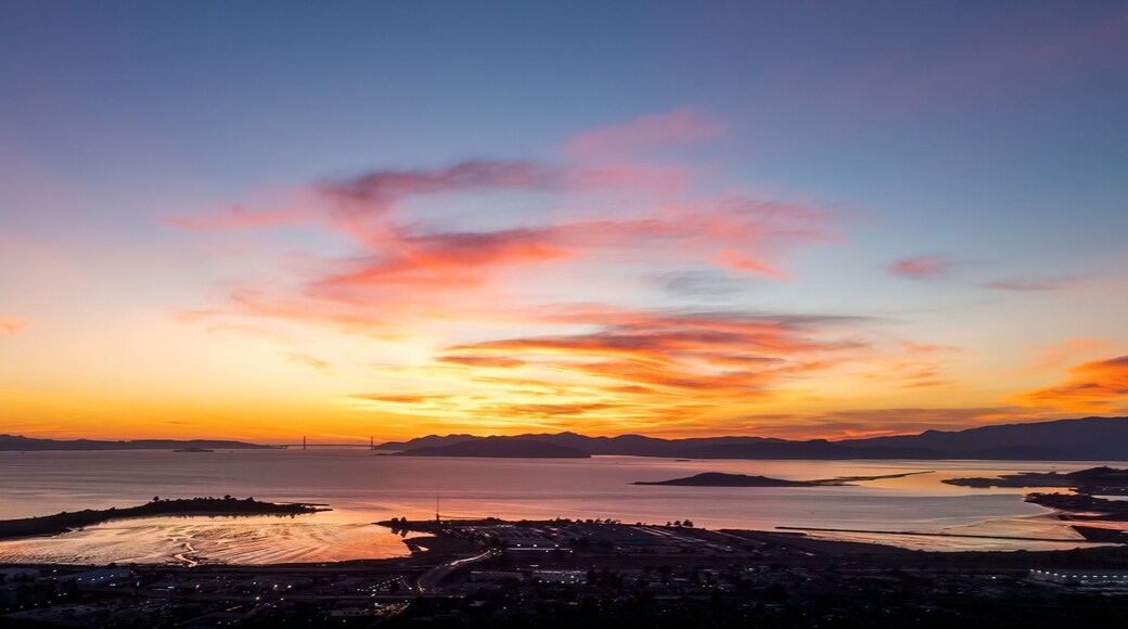 A sunset over a coastal city with a bridge, mountains, and a calm sea.