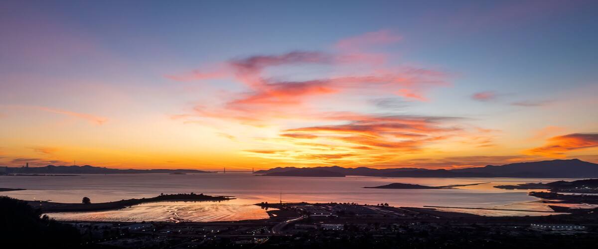 A sunset over a coastal city with a bridge, mountains, and a calm sea.