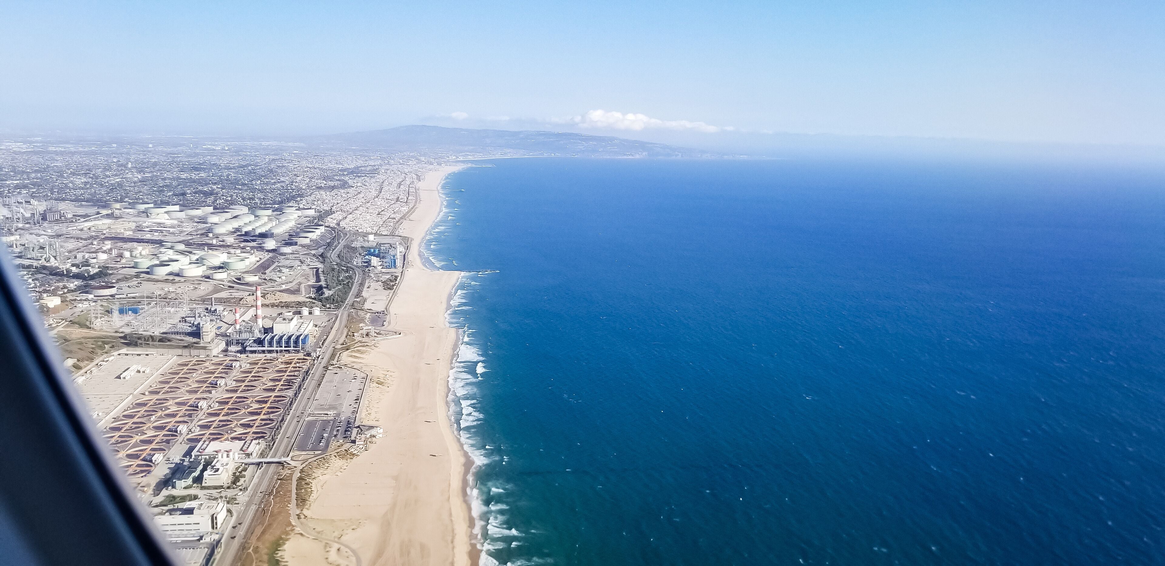 Vista aérea de uma cidade costeira com uma praia extensa, área industrial e mar azul cristalino.