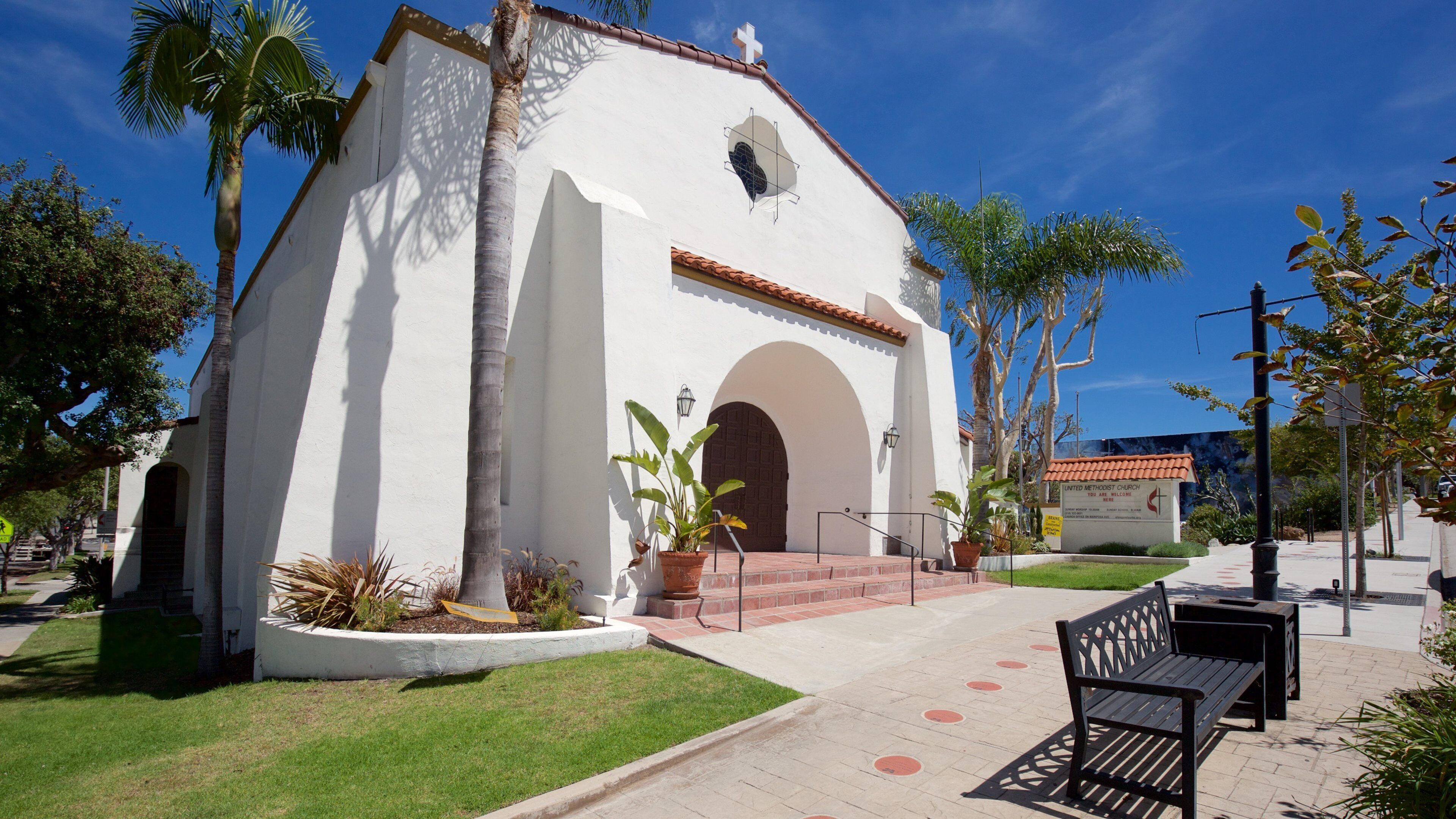 A white church with a red-tiled roof, surrounded by palm trees and a paved walkway.