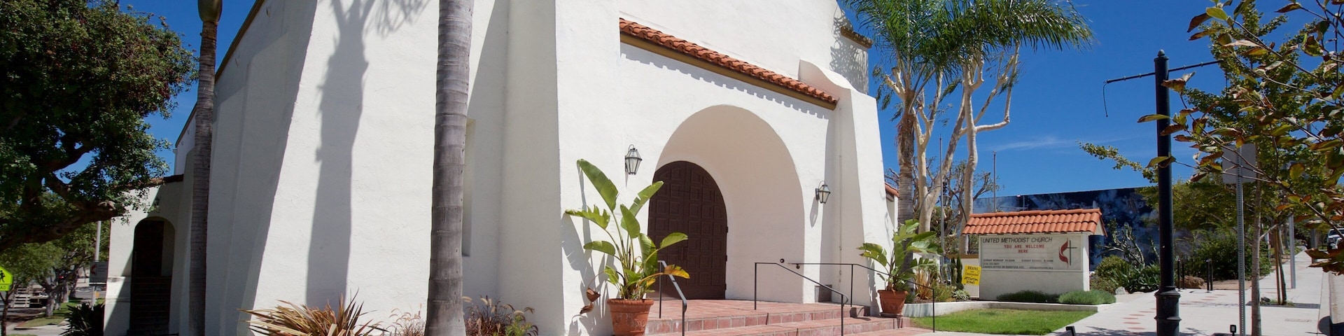 A white church with a red-tiled roof, surrounded by palm trees and a paved walkway.