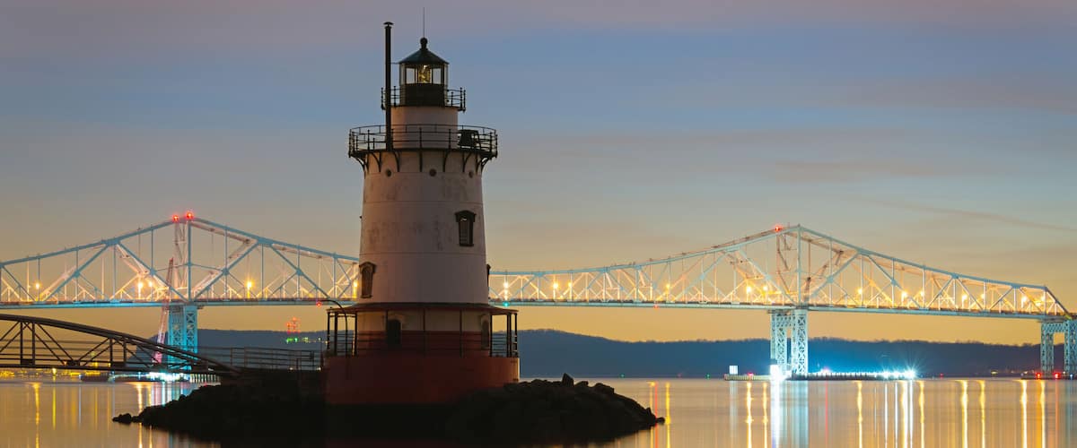 A lighthouse on a small island with a bridge in the background.
