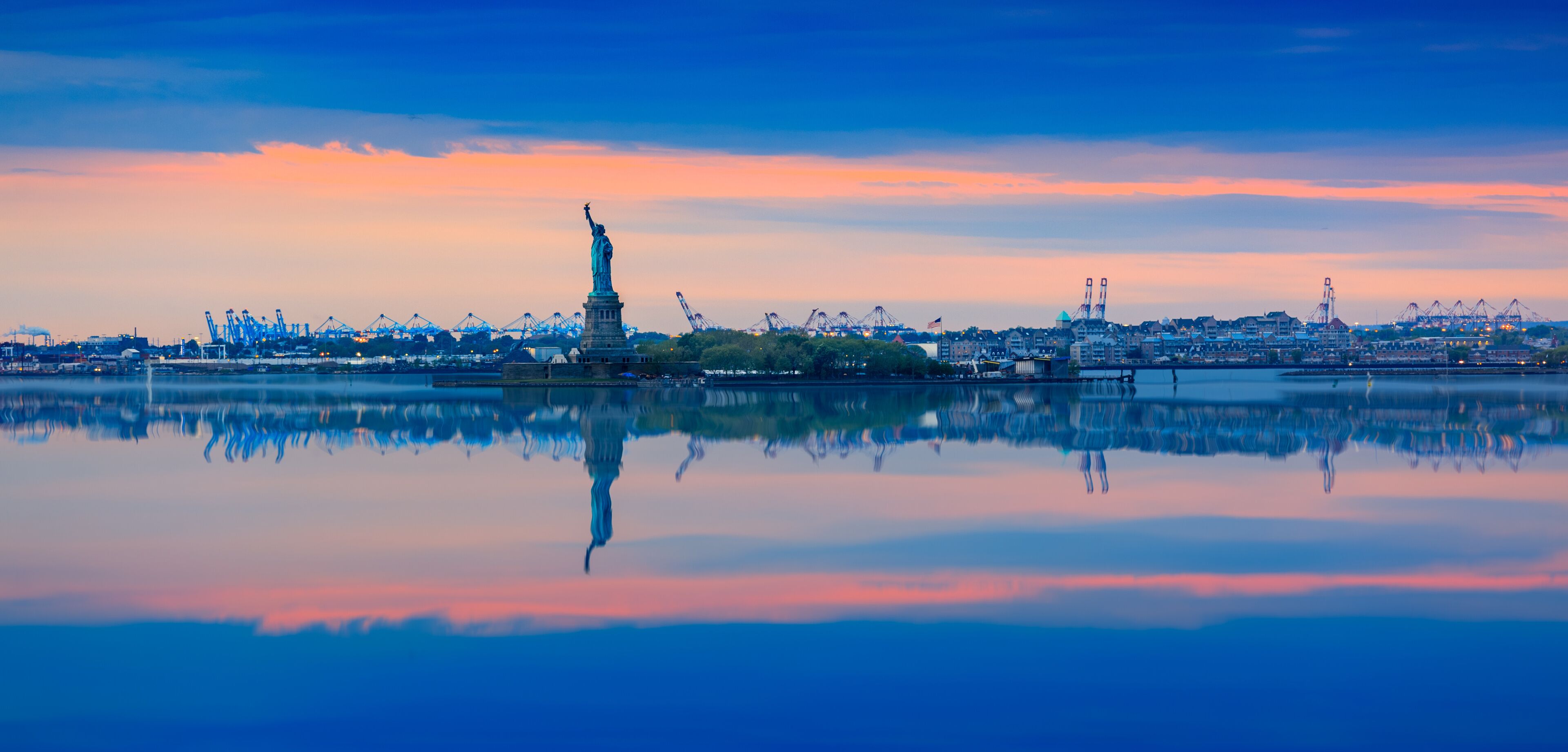 The Statue of Liberty reflected in calm water, with a city skyline and cranes in the background.