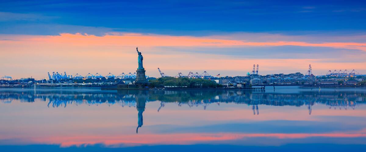 The Statue of Liberty reflected in calm water, with a city skyline and cranes in the background.