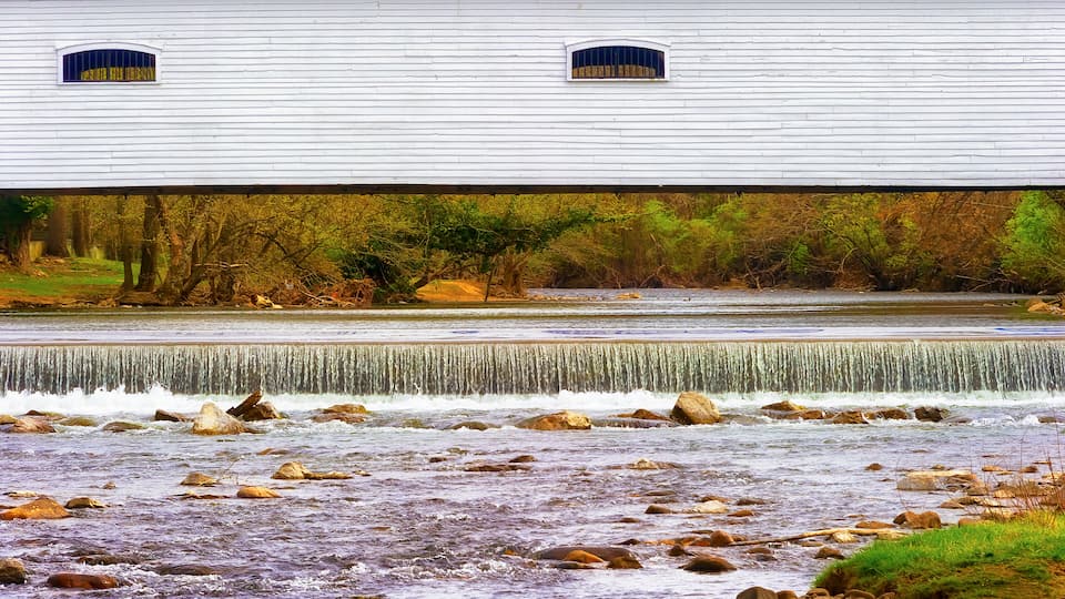 A white building with two windows, a river with rocks, and a waterfall.