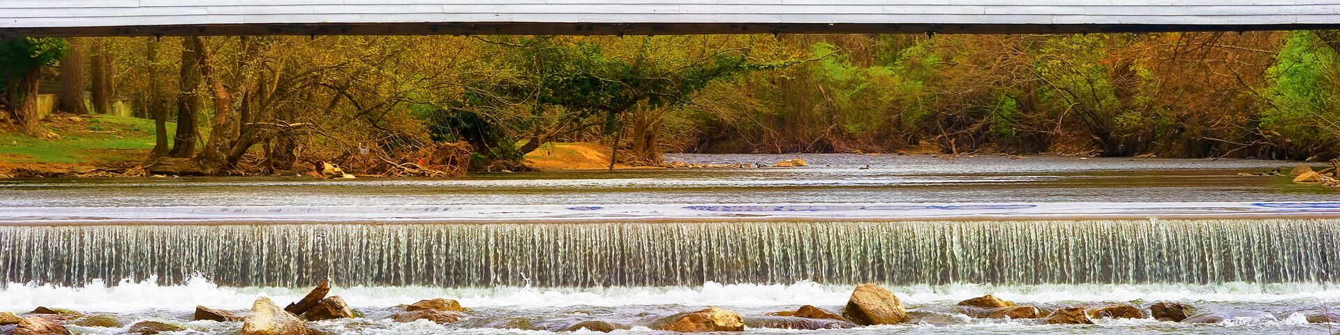 Un edificio blanco con dos ventanas, un río con rocas y una cascada.