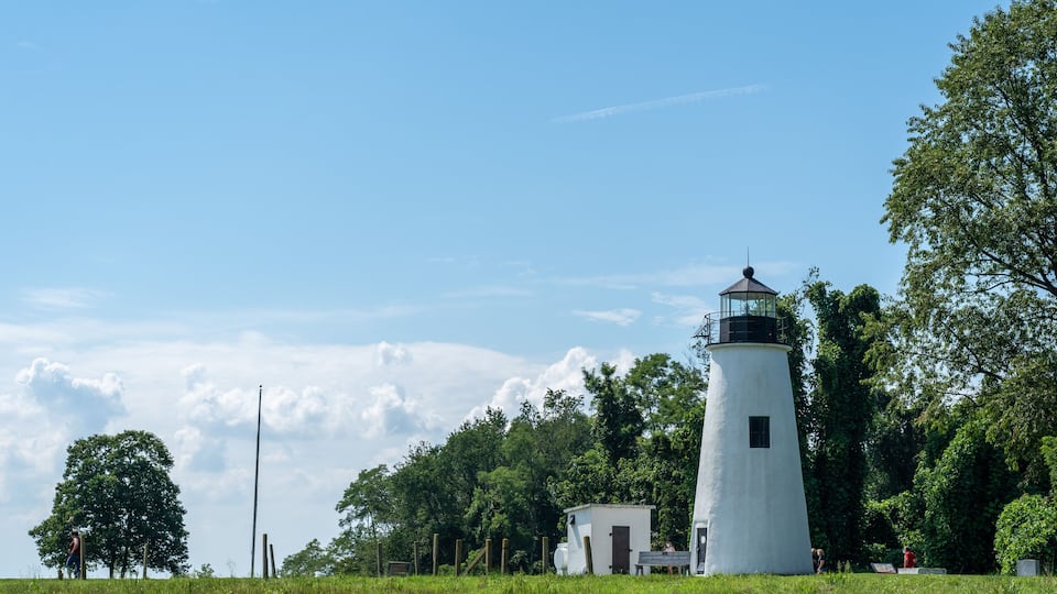A white lighthouse with a black top, surrounded by lush green trees and a clear blue sky.
