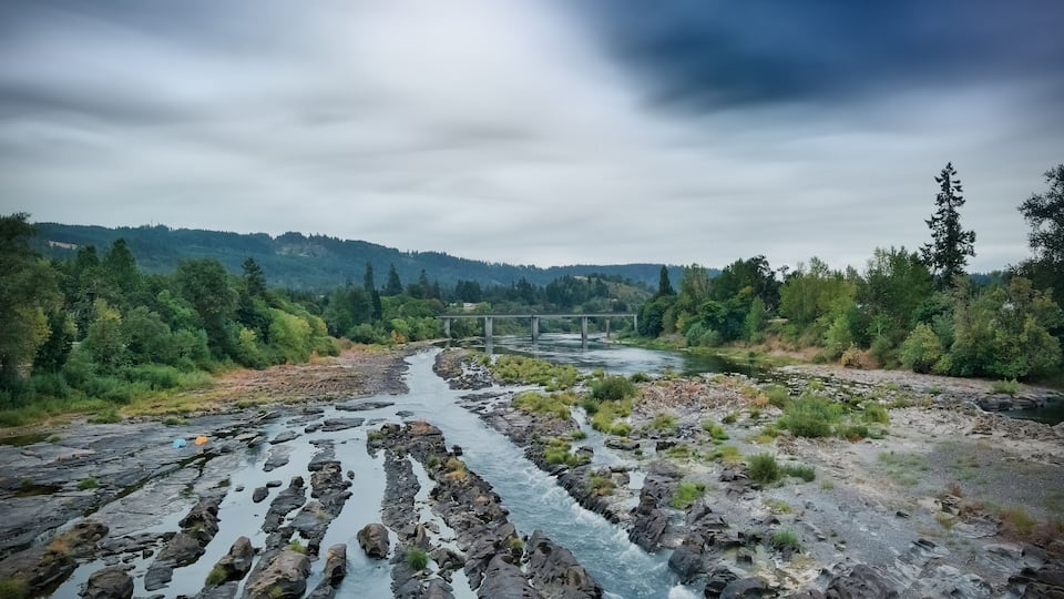 A river flowing through a rocky landscape with a bridge in the distance and dense forest on either side.