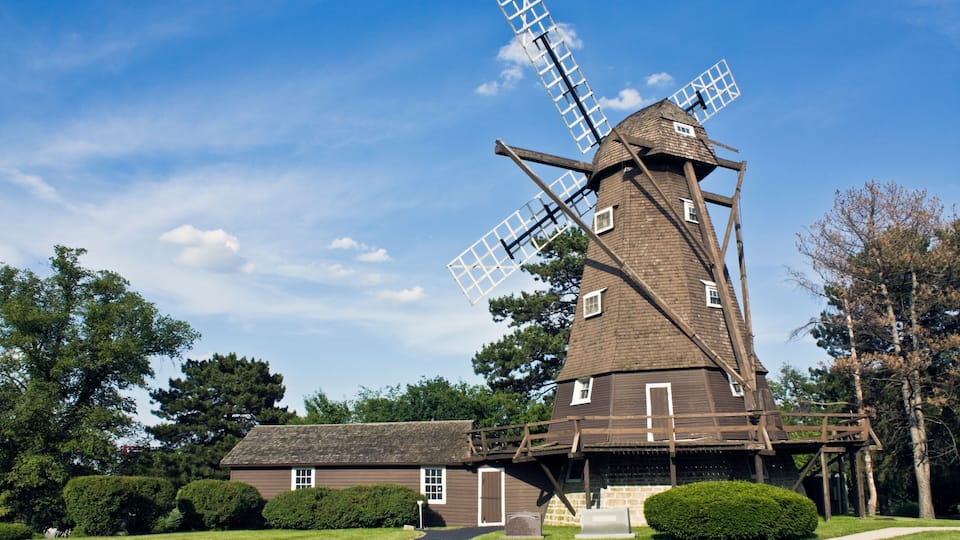 A traditional windmill with a wooden structure and large sails, surrounded by a well-maintained lawn and trees.