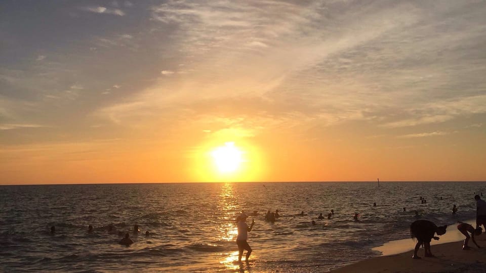 A sunset over a beach with people swimming and walking along the shore.