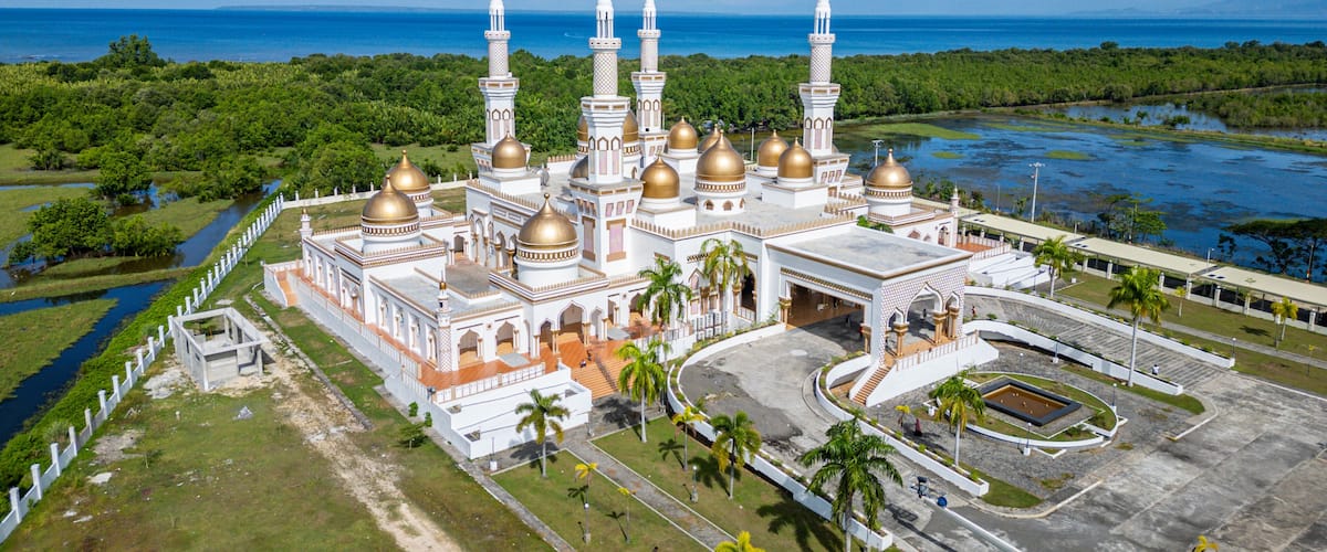 A large white mosque with golden domes and minarets, surrounded by lush greenery and a body of water.