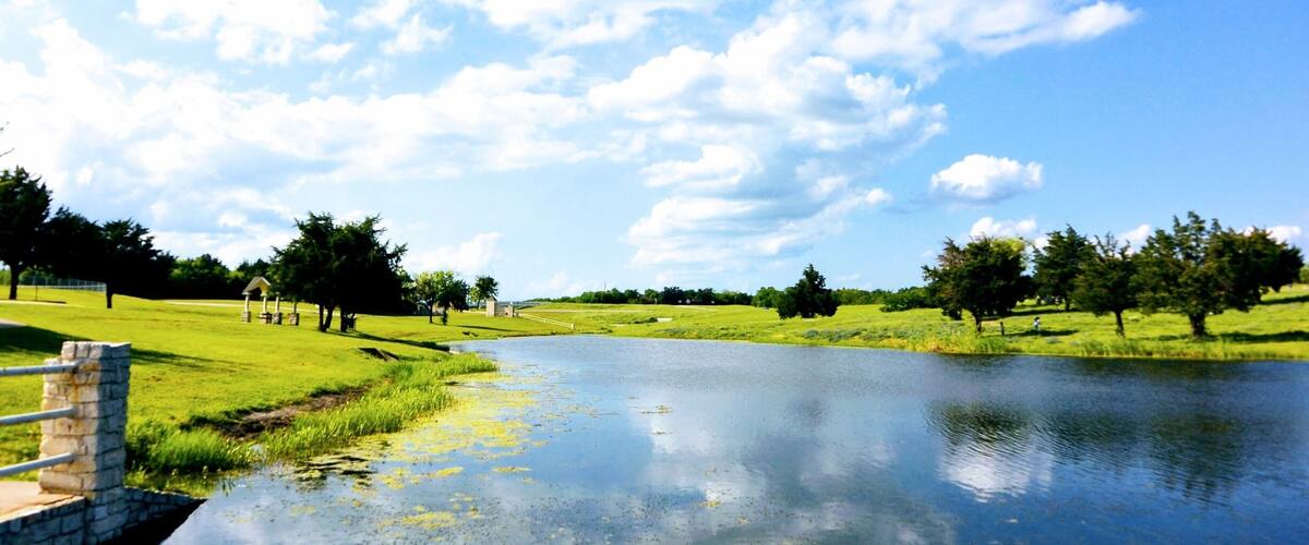 A serene landscape with a calm river, grassy fields, scattered trees, and a stone bridge.