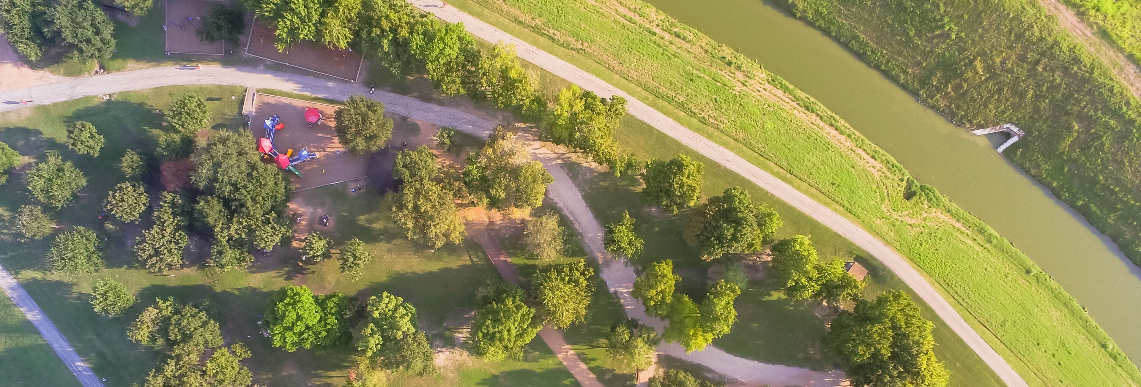 An aerial view of a park with playground equipment, a dirt path, and a river.