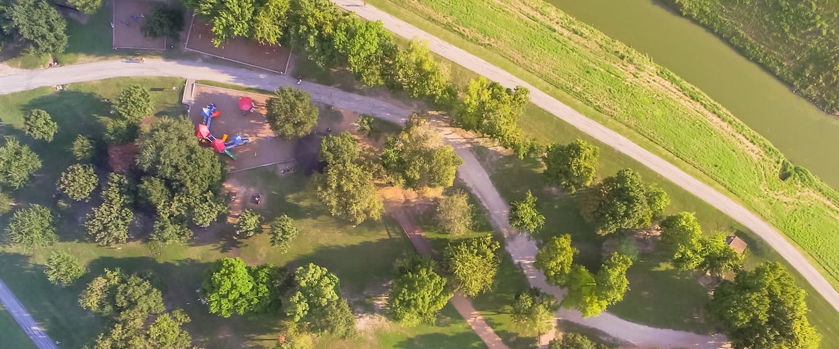 An aerial view of a park with playground equipment, a dirt path, and a river.