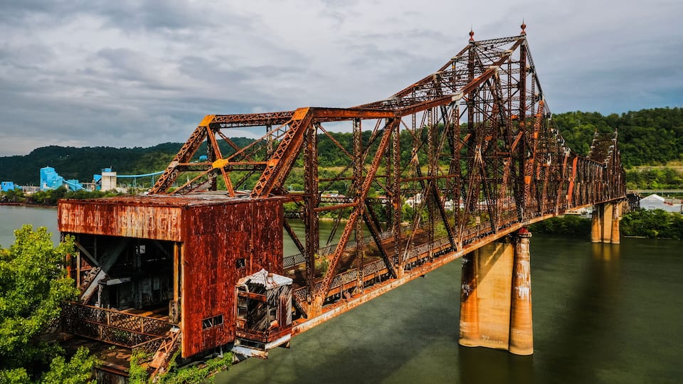 A rusted steel bridge over a river with a forested hillside in the background.